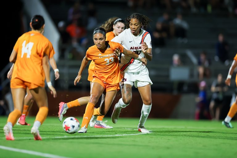Two soccer players compete for the ball during a nighttime match, with one in an orange jersey and another in a white jersey.