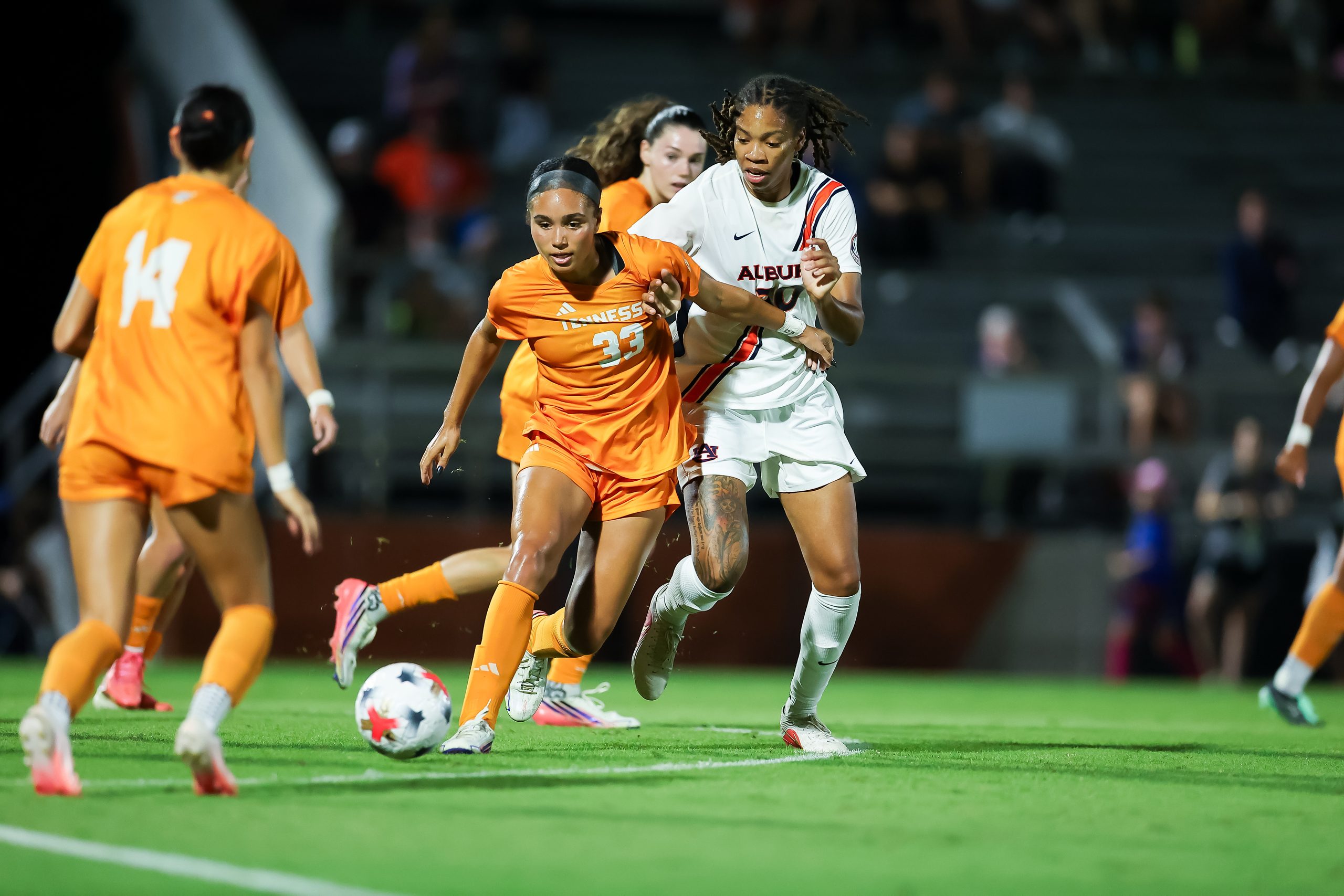 Two soccer players compete for the ball during a nighttime match, with one in an orange jersey and another in a white jersey.
